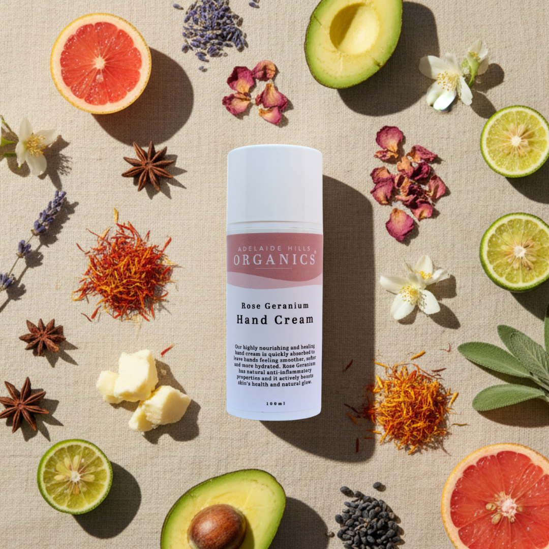 Rose Geranium Hand Cream surrounded by fruits, flowers, and leaves on a textured surface